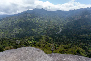 View from a rocky ledge overlooking a lush, green mountain valley, with dense forests, a winding river, and distant mountain ridges under a partly cloudy sky.
