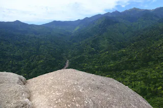 A large rock in the foreground overlooks a lush, green forest valley with mountains rising in the distance under a partly cloudy sky.