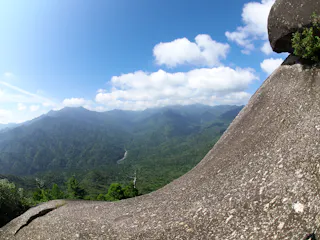A large, smooth rock slopes down toward a lush green forest with mountains in the background under a blue sky with scattered clouds.