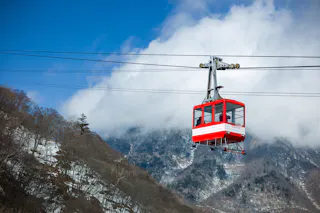 A red and white cable car travels above a snowy mountain landscape with clouds and blue sky in the background.