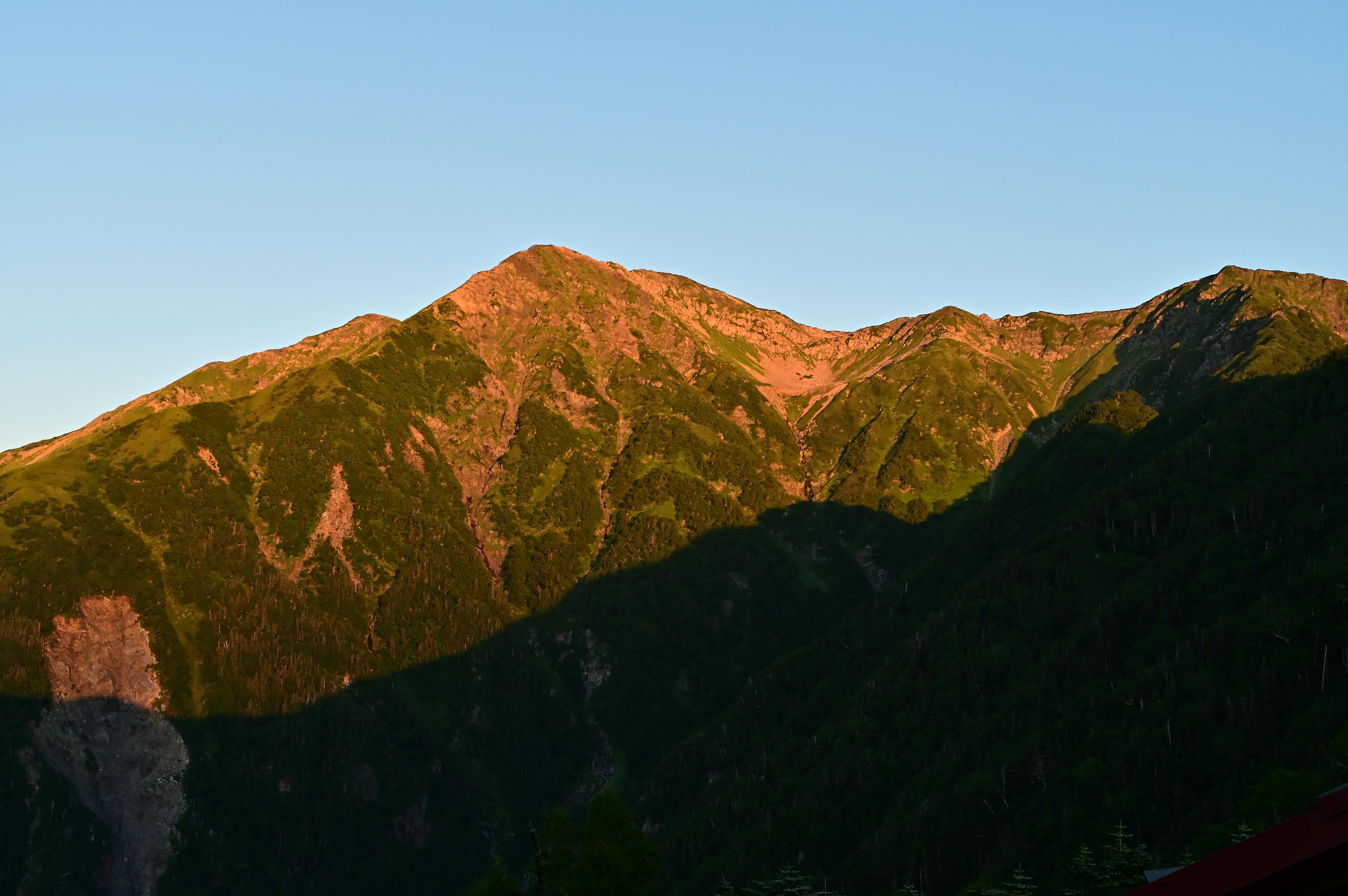 A mountain range bathed in warm sunlight, with peaks tinged in orange and green forests covering the slopes. A deep shadow stretches across the lower part of the mountains under a clear blue sky.