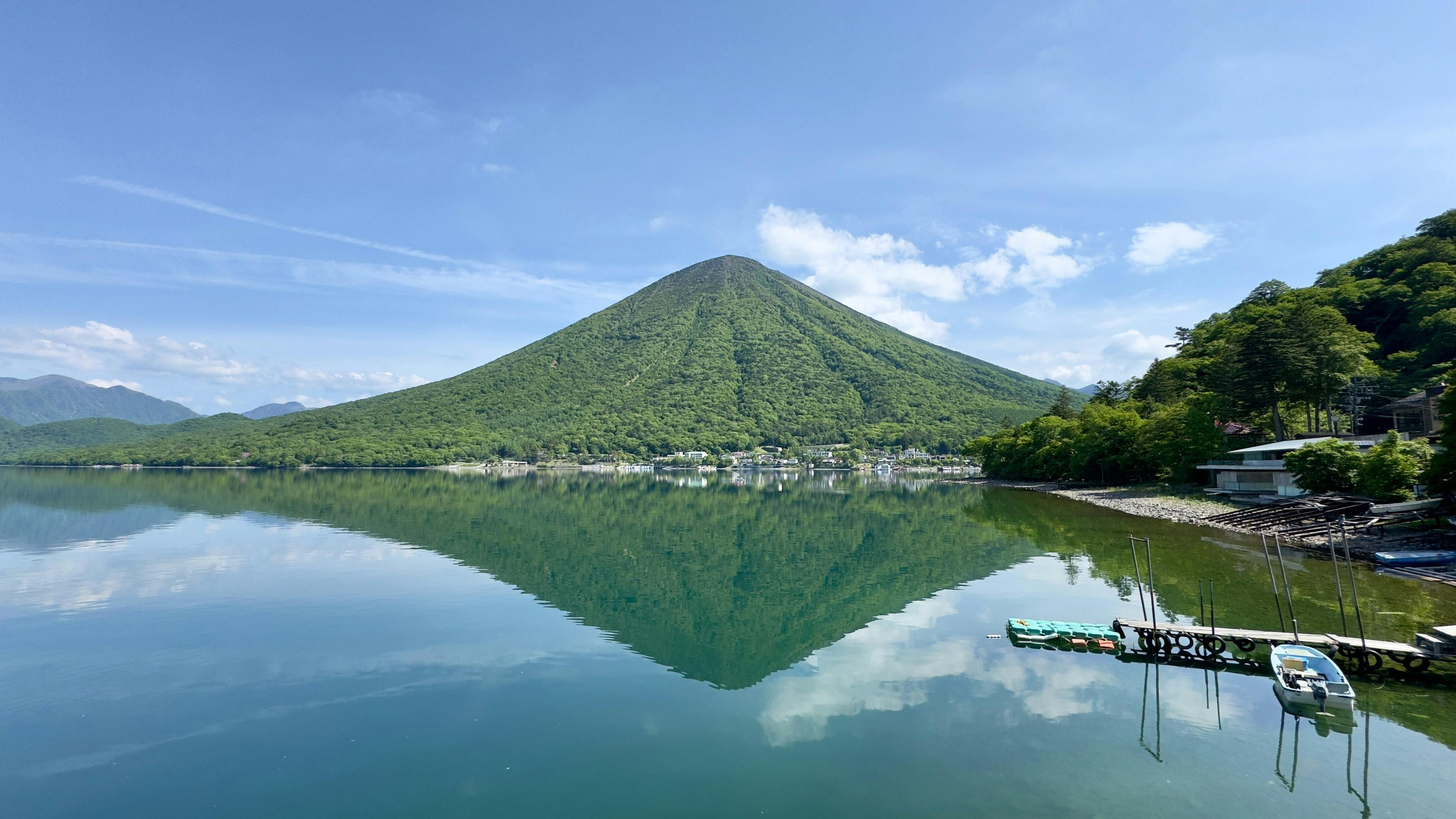A green, cone-shaped mountain is reflected in a calm lake under a blue sky with scattered clouds. Trees and small buildings line the shore, and several boats are docked at a wooden pier on the right.