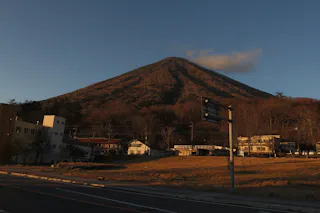 A mountain covered in brownish foliage is lit by warm sunlight at sunset, with a few buildings and houses at its base and a clear sky above. A road and street signs are visible in the foreground.