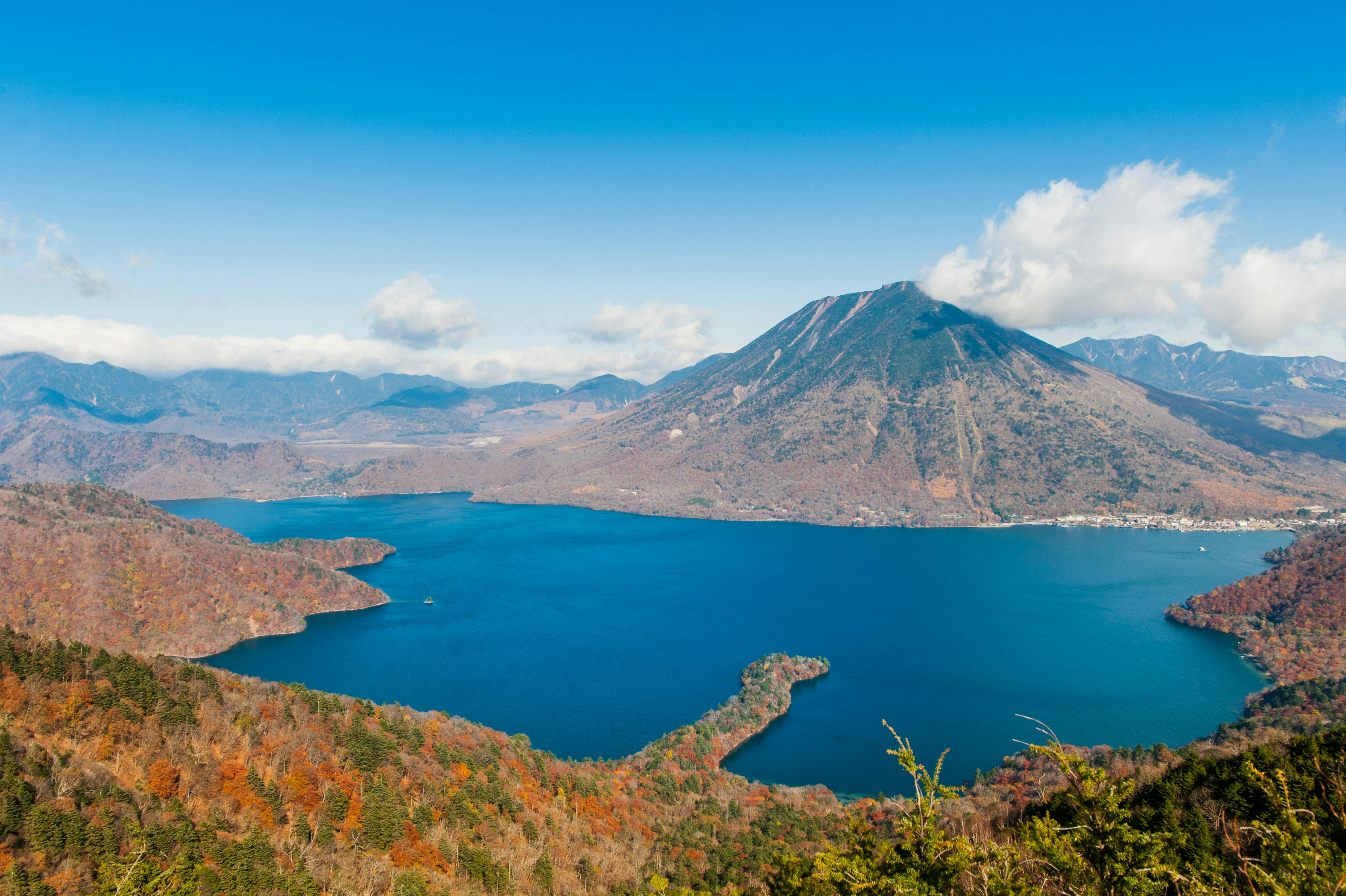 A panoramic view of a deep blue lake surrounded by forested hills with autumn foliage, and a tall mountain in the background under a clear blue sky with scattered clouds.