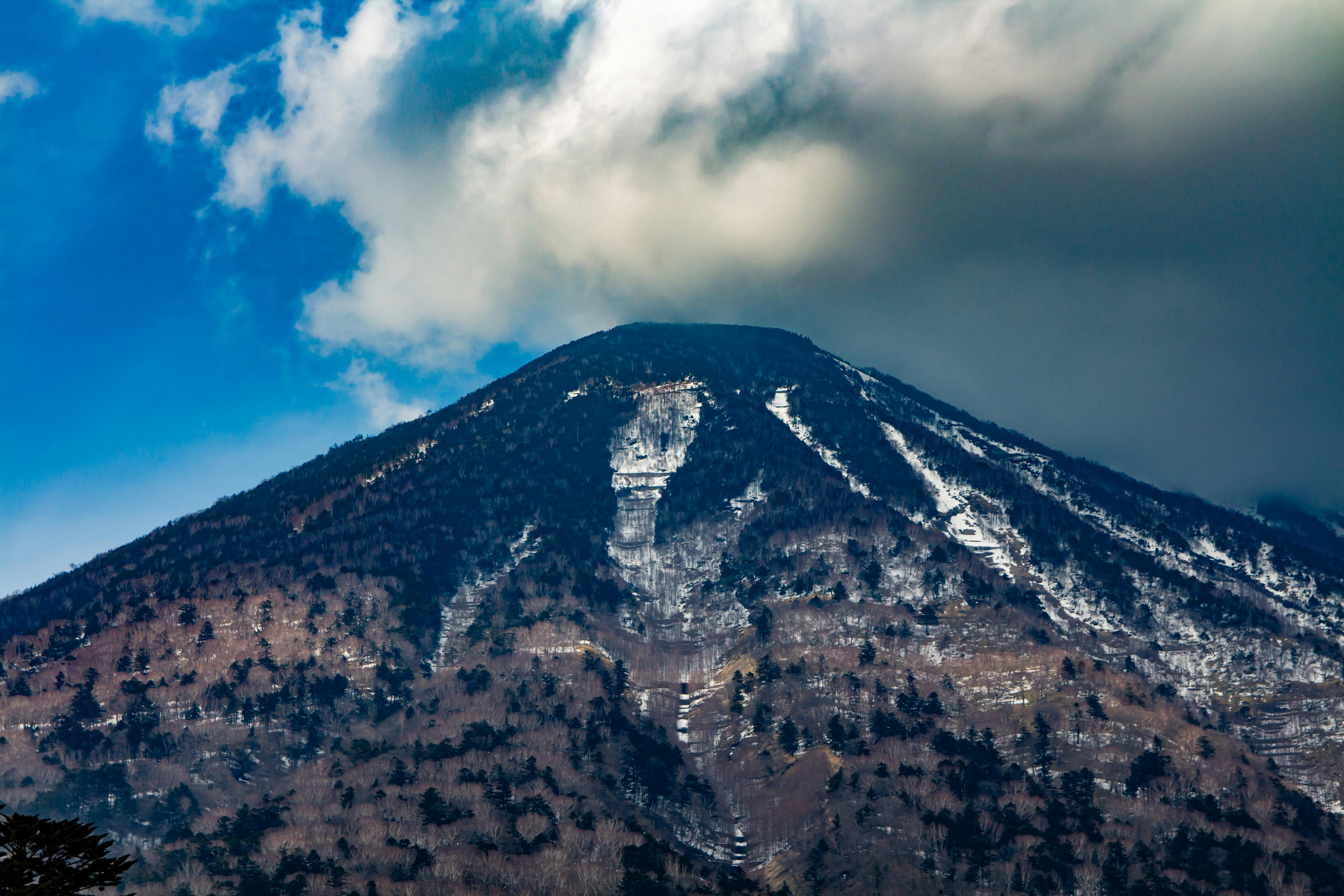 A mountain with sparse trees and patches of snow on its slopes stands beneath a partly cloudy sky with a mix of blue and gray clouds.