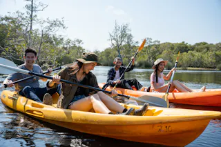 Four young adults paddle kayaks on a calm river, smiling and enjoying the outdoors. Green trees line the riverbank under a clear sky. Two people are in each kayak, wearing casual clothes and hats.