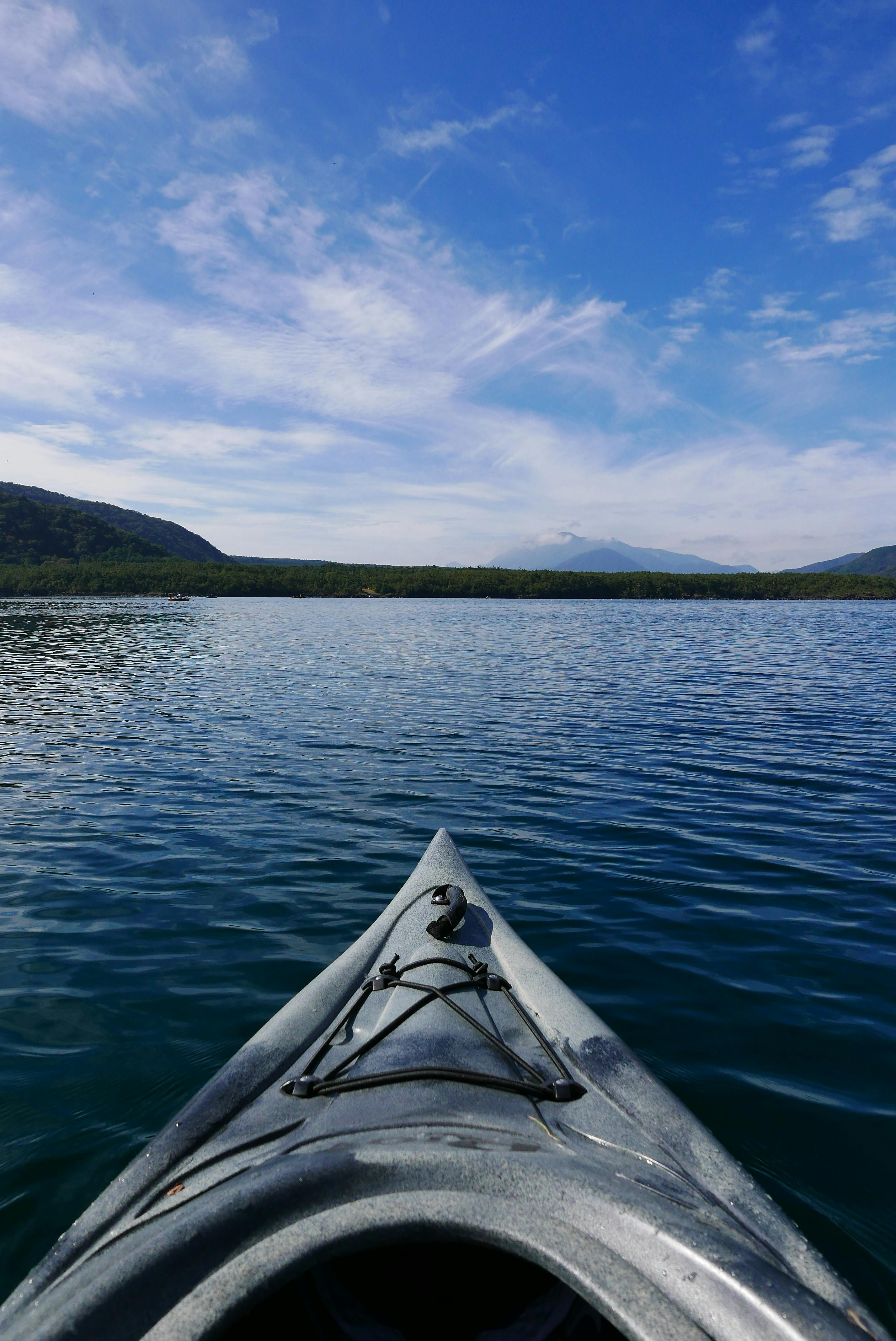 A kayak's bow glides over calm, blue lake water under a bright sky with scattered clouds; green hills and distant mountains line the horizon.