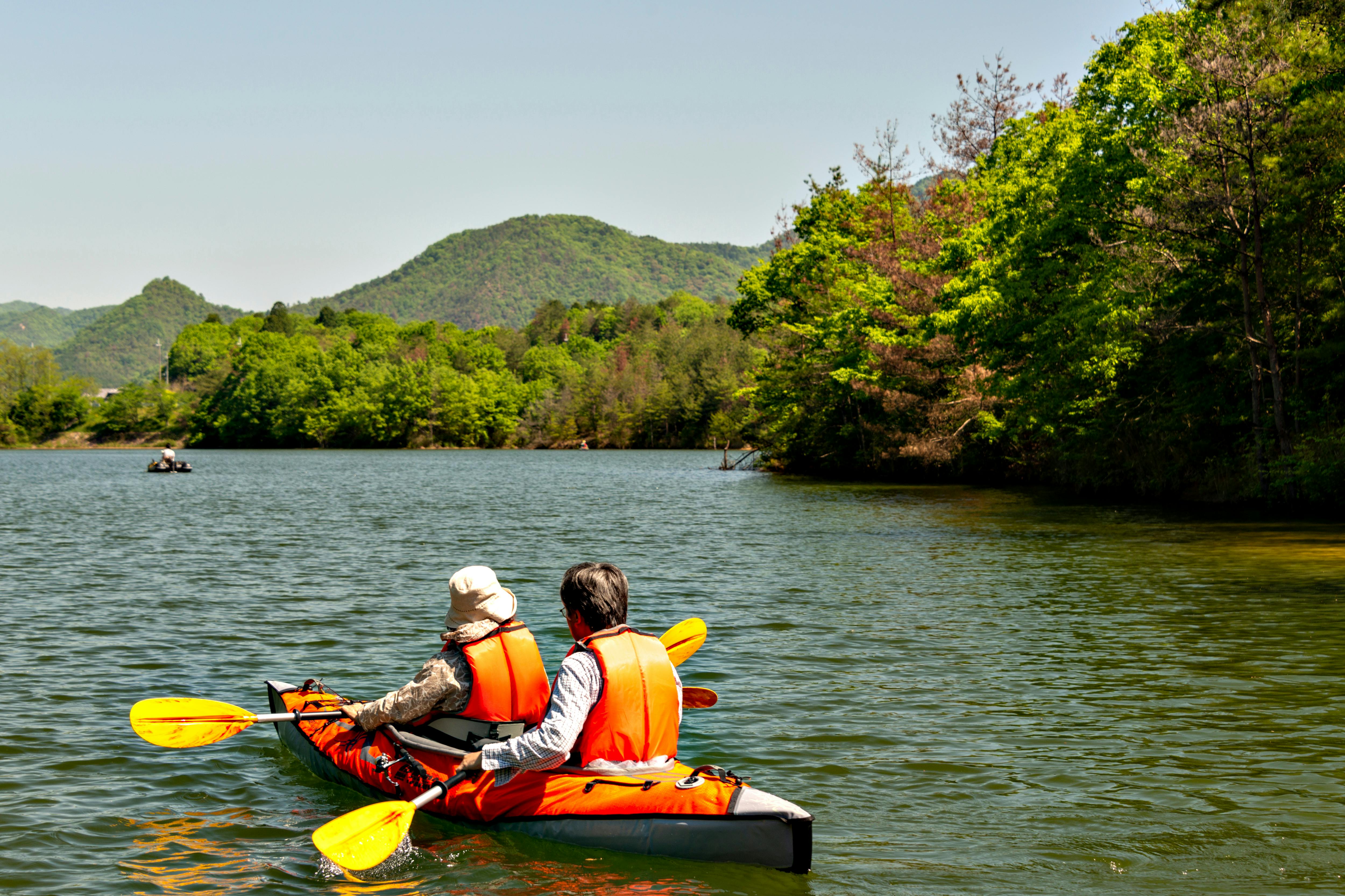 Two people wearing orange life jackets paddle a kayak on a calm lake surrounded by lush green trees and distant hills under a clear sky.