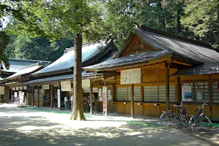 Traditional Japanese wooden shrine buildings surrounded by trees, with informational signs and bicycles parked nearby; a few people are visible near the entrance.