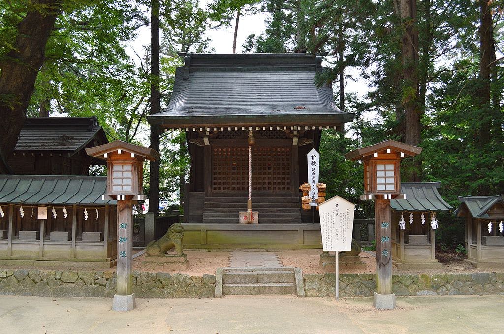 A small traditional Japanese Shinto shrine stands surrounded by tall trees, with two wooden lanterns at the entrance and a white signboard to the right of the stone steps leading up to the shrine.