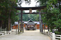 A traditional Japanese Shinto shrine entrance with two large wooden torii gates, surrounded by tall trees. Beyond the gates, a wooden building with a tiled roof stands in a tranquil, natural setting.