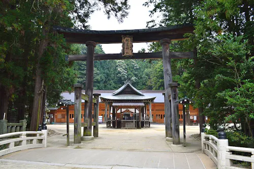 A traditional Japanese Shinto shrine entrance with two large wooden torii gates, surrounded by tall trees. Beyond the gates, a wooden building with a tiled roof stands in a tranquil, natural setting.