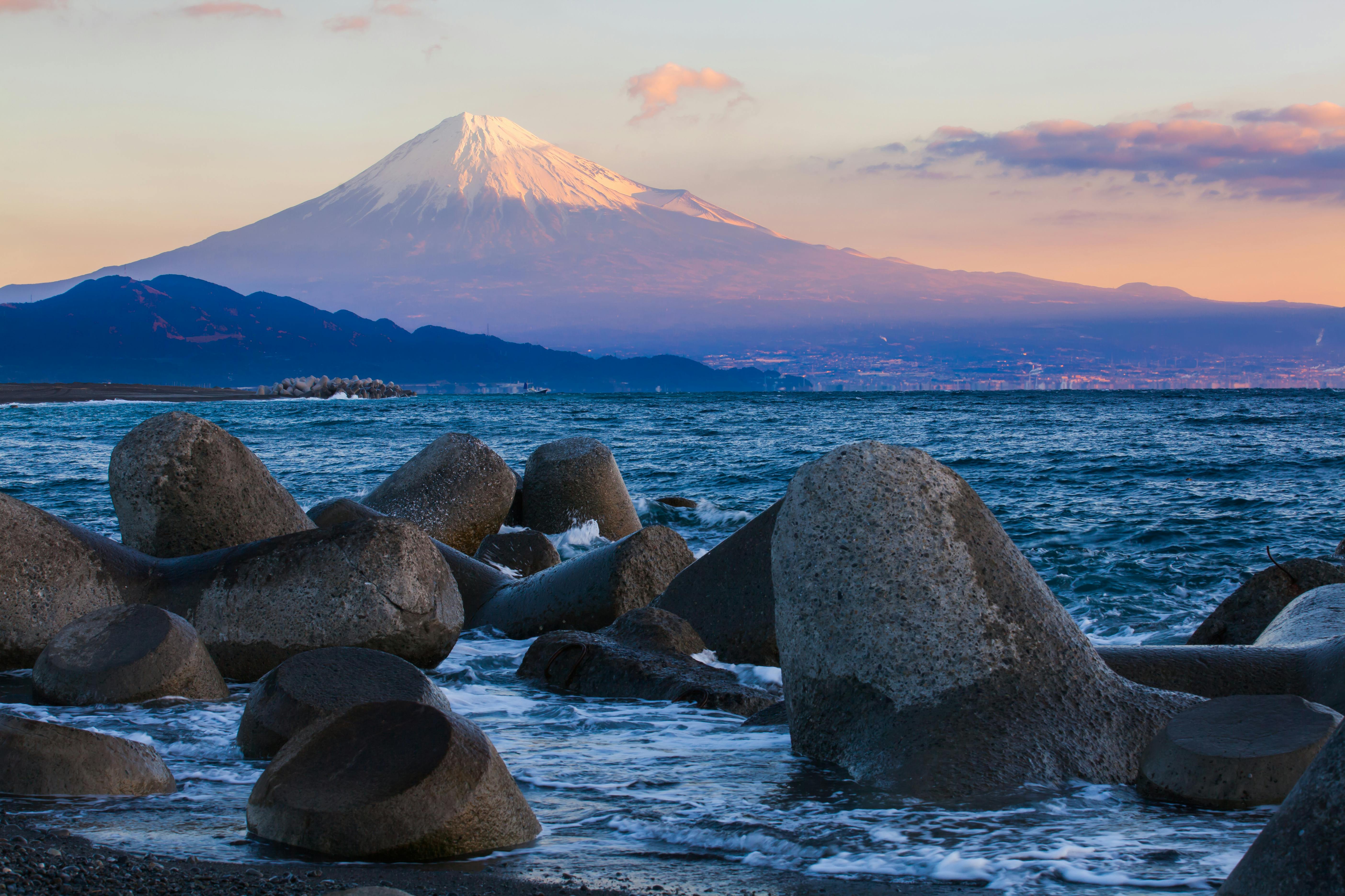 Large concrete tetrapods line the shore with ocean waves washing against them, while Mount Fuji rises in the background, its snow-capped peak illuminated by the soft light of sunrise or sunset.