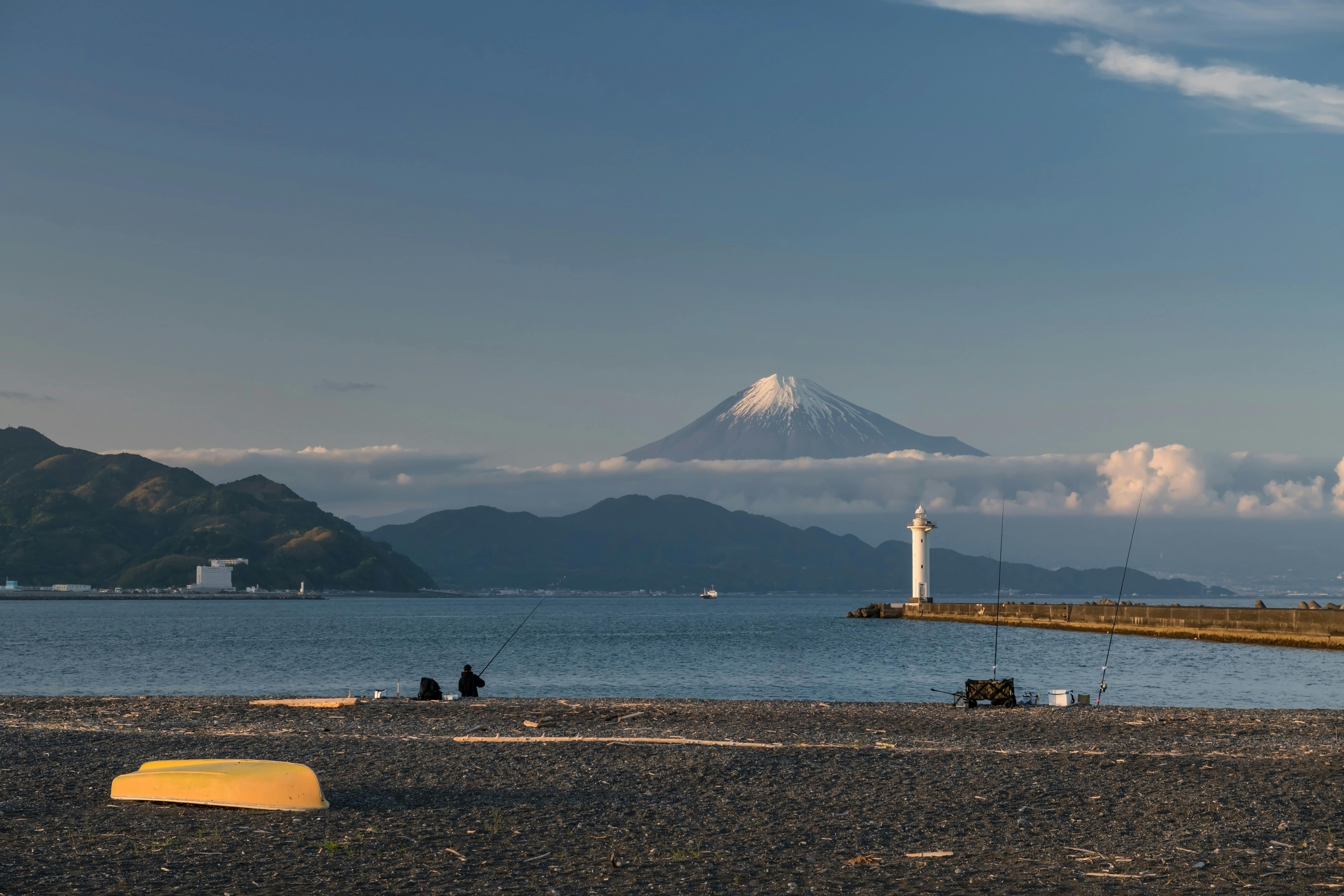 Two people fish on a pebble beach near a yellow boat, with a harbor, a white lighthouse, calm water, and Mount Fuji in the background under a blue sky with clouds.
