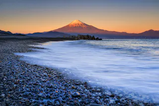 A serene shoreline with gentle waves, pebbled beach, and Mount Fuji in the distance, its snow-capped peak glowing as the sun sets, casting an orange hue across the sky.