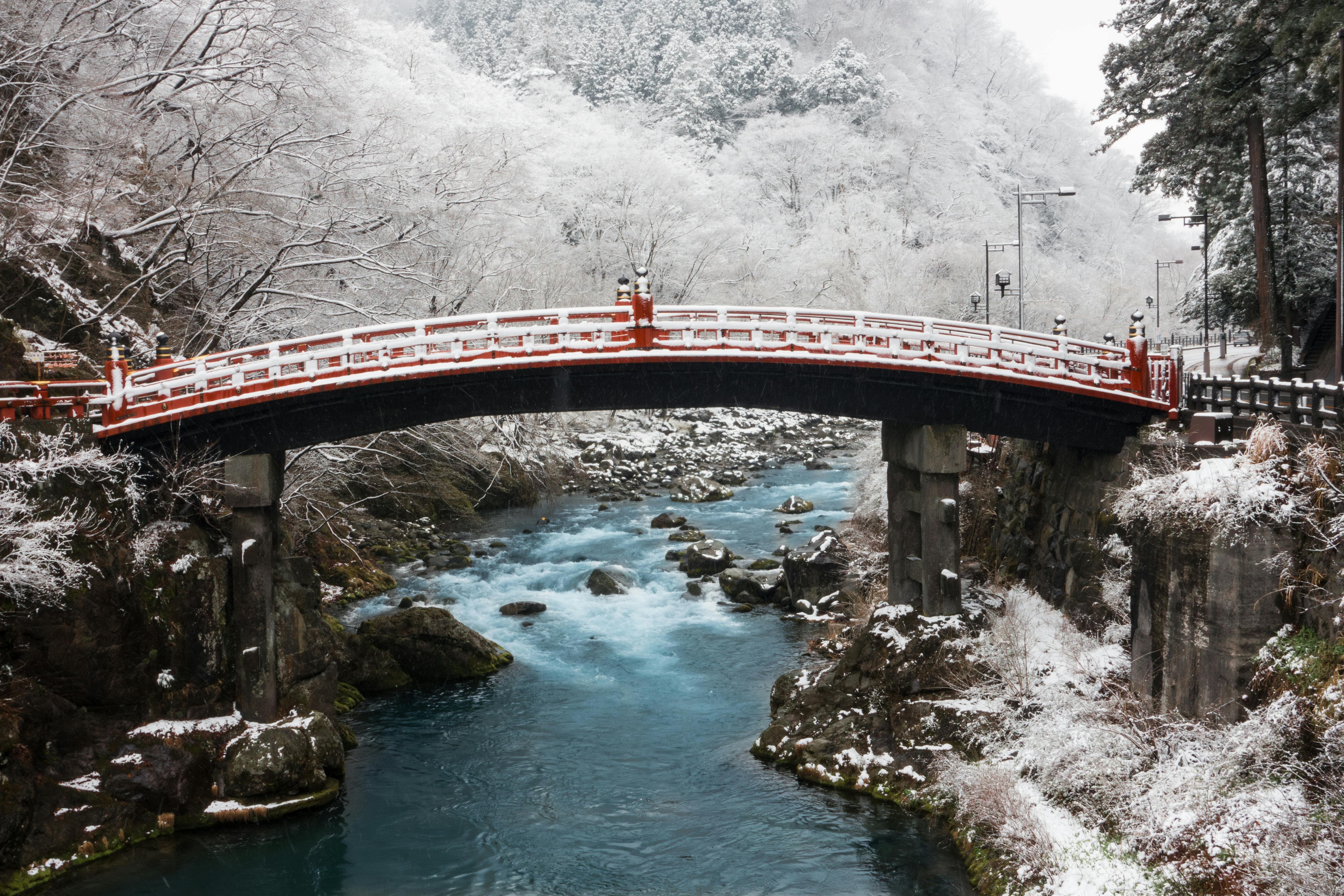 A red and white arched bridge spans a turquoise river surrounded by snow-covered trees and rocky banks in a serene winter landscape.