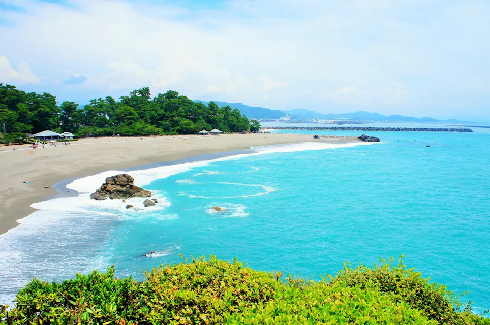 Scenic view of a sandy beach with clear turquoise water, surrounded by lush green trees. Several large rocks are scattered along the shoreline. The sky is lightly clouded, and a distant mountain range is faintly visible in the background.