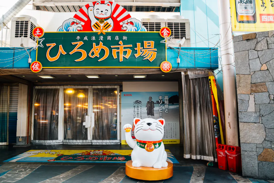 An entrance to a Japanese market with colorful signage reading "ひろめ市場" (Hirome Market). Above the door is a large waving lucky cat, and a matching statue stands in front, wearing a bib and a bell. Stone walls and vending machines are on either side of the entrance.