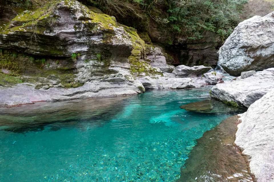 A serene natural pool with crystal-clear turquoise water surrounded by moss-covered rocks and lush greenery. The water is calm, reflecting the surrounding rocks and foliage, creating a peaceful and refreshing atmosphere in this untouched, scenic location.