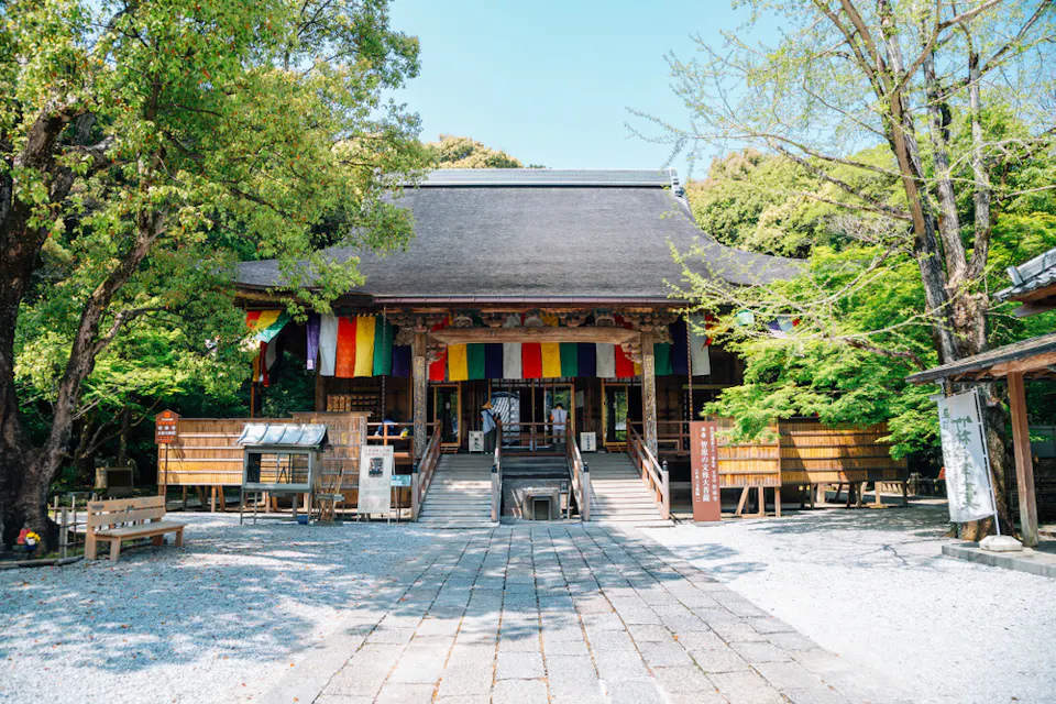 A traditional Japanese temple with a steep, thatched roof and colorful prayer flags hanging from the eaves. Stairs lead up to the entrance, flanked by lush green trees. Several wooden plaques and small structures are positioned around the temple grounds.