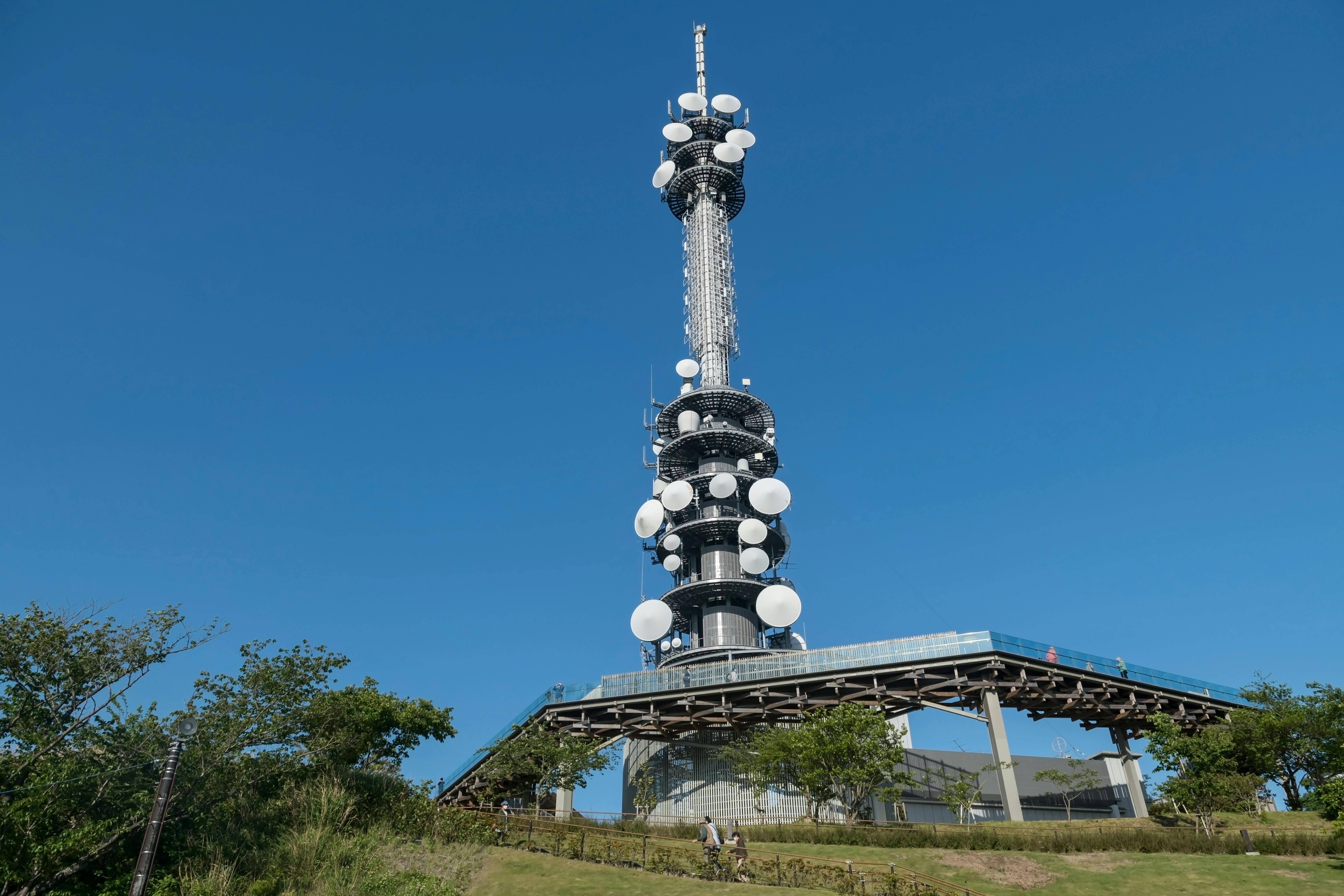 A tall telecommunications tower with multiple circular antennas stands on a grassy hill under a clear blue sky, surrounded by trees and a raised platform at its base.