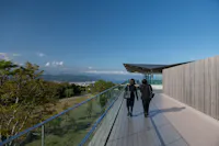 Two people in black clothing walk along a glass-walled terrace with a wooden deck. The building has a modern design. A scenic view of a park and the sea is visible under a clear blue sky.