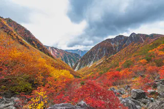 A scenic mountain valley covered in vibrant autumn foliage with red, orange, and yellow trees, rocky terrain in the foreground, and dramatic, cloudy sky overhead.