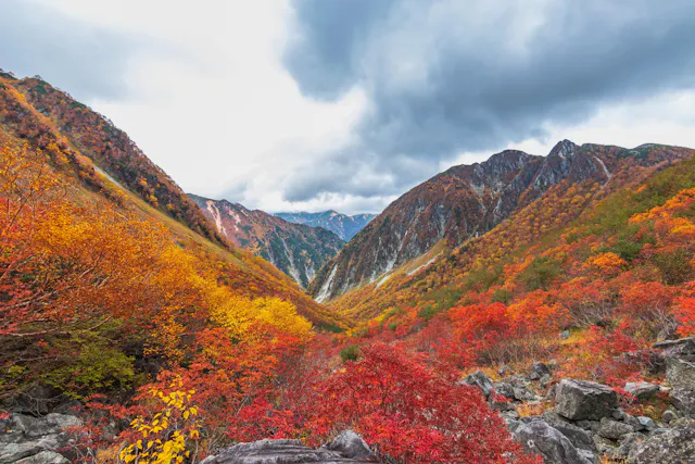 A scenic mountain valley covered in vibrant autumn foliage with red, orange, and yellow trees, rocky terrain in the foreground, and dramatic, cloudy sky overhead.