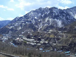 A mountain village with scattered buildings sits at the base of tall, rugged mountains partly covered in snow, under a partly cloudy sky. Sparse trees cover the lower slopes and some rooftops are visible.