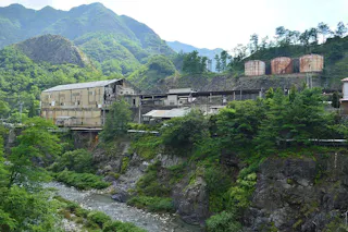 An old industrial building and two large rusted storage tanks sit on a rocky hillside surrounded by green trees and mountains, with a narrow river flowing below.