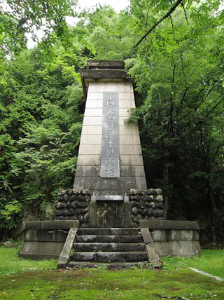 A tall stone monument with Chinese inscriptions stands in a lush, green forest. The monument sits atop a stepped platform, surrounded by trees and dense foliage.