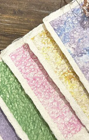 Washi Papermaking Experience A set of handmade textured cards with pastel colors—purple, green, pink, yellow, and blue—arranged in a fan shape on a wooden surface, with dried purple flowers beside them.