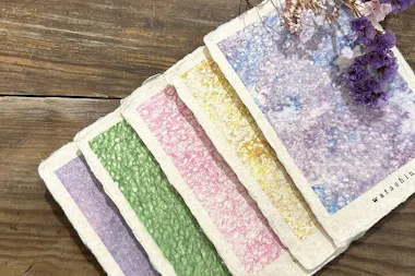 A set of handmade textured cards with pastel colors—purple, green, pink, yellow, and blue—arranged in a fan shape on a wooden surface, with dried purple flowers beside them.
