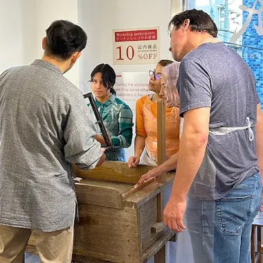 Washi Papermaking Experience Four people stand around a wooden box in a workshop, focusing on a hands-on activity. Two people wear aprons, and sunlight streams through large windows with blue writing and signs visible outside.