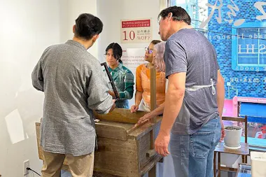 Four people stand around a wooden box in a workshop, focusing on a hands-on activity. Two people wear aprons, and sunlight streams through large windows with blue writing and signs visible outside.