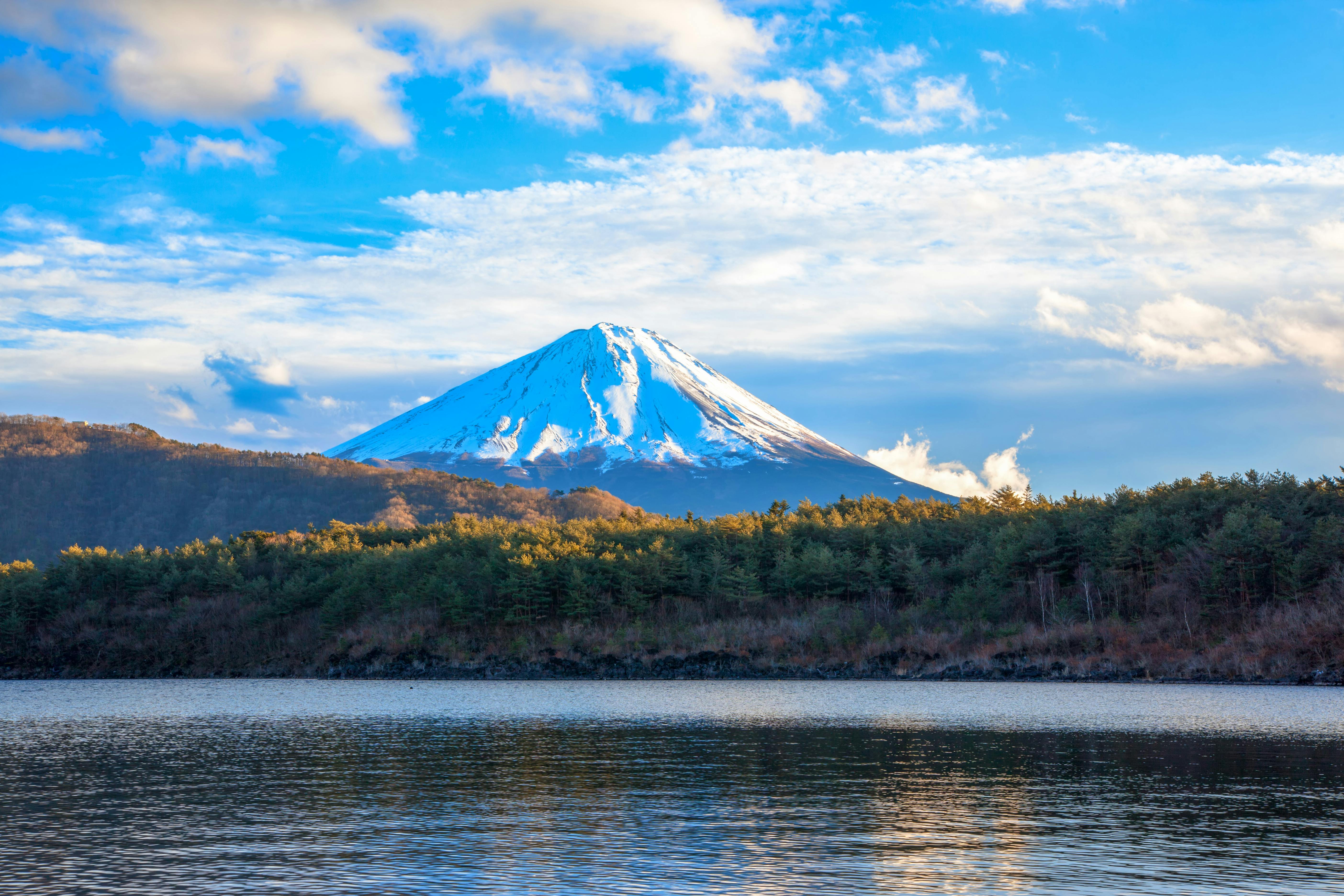 Snow-capped mountain with a forest at its base, reflected in a calm lake under a partly cloudy blue sky.