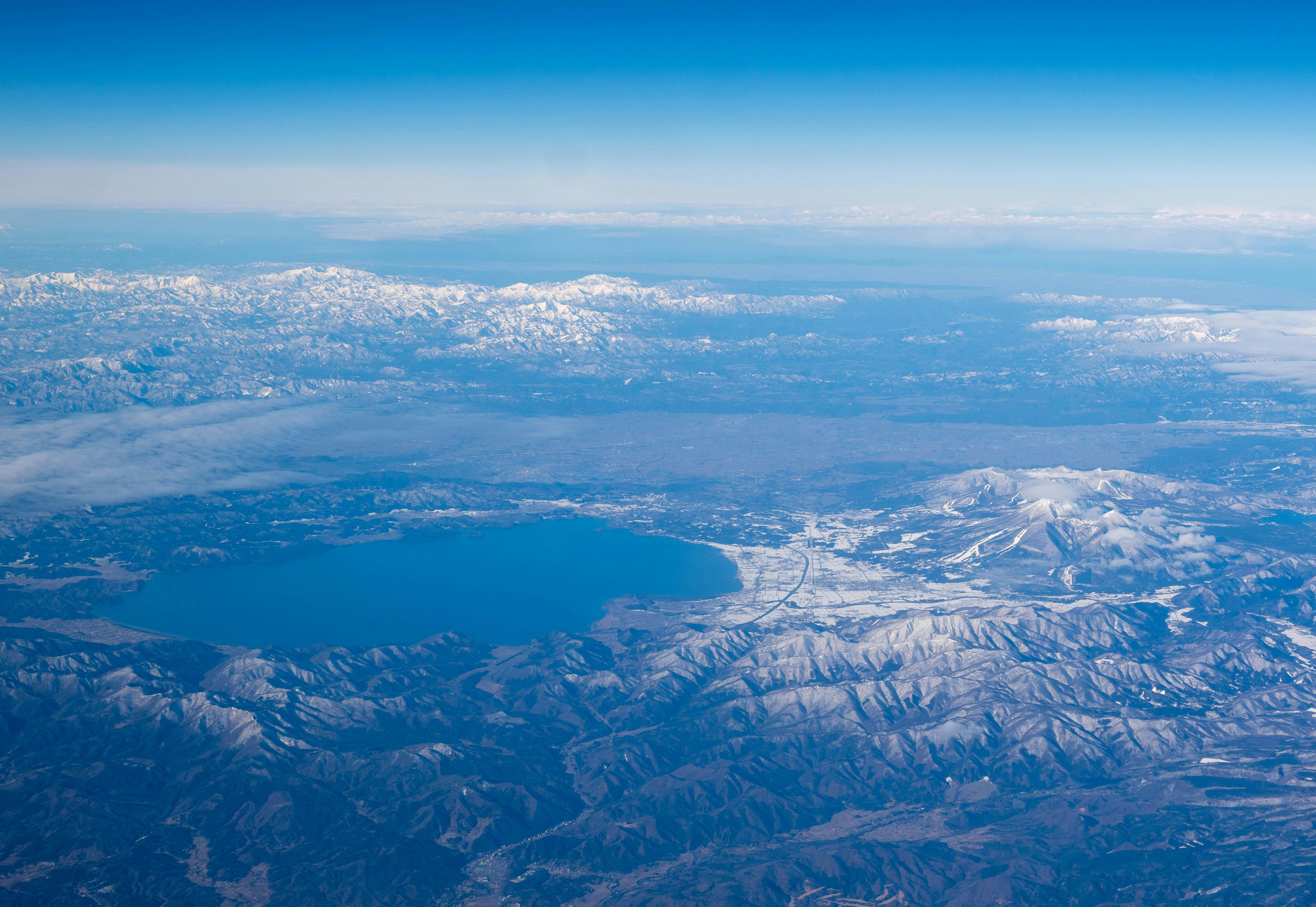 Aerial view of a large blue lake surrounded by snow-capped mountains and valleys under a clear blue sky, with distant mountain ranges visible on the horizon.