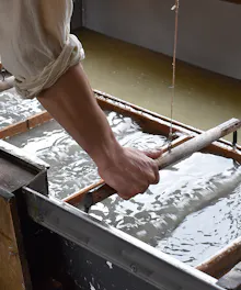 Washi Papermaking A person uses a wooden frame to make paper by hand, dipping it into a vat of water and pulp. The person’s sleeves are rolled up, and the surface is covered with wet pulp.