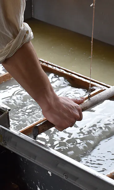 Washi Papermaking A person uses a wooden frame to make paper by hand, dipping it into a vat of water and pulp. The person’s sleeves are rolled up, and the surface is covered with wet pulp.