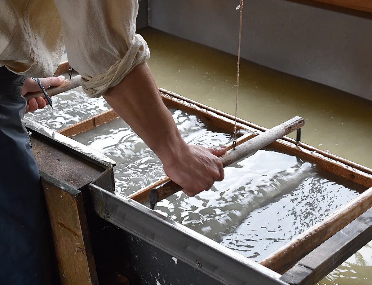 Washi Papermaking A person uses a wooden frame to make paper by hand, dipping it into a vat of water and pulp. The person’s sleeves are rolled up, and the surface is covered with wet pulp.