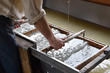 A person uses a wooden frame to make paper by hand, dipping it into a vat of water and pulp. The person’s sleeves are rolled up, and the surface is covered with wet pulp.