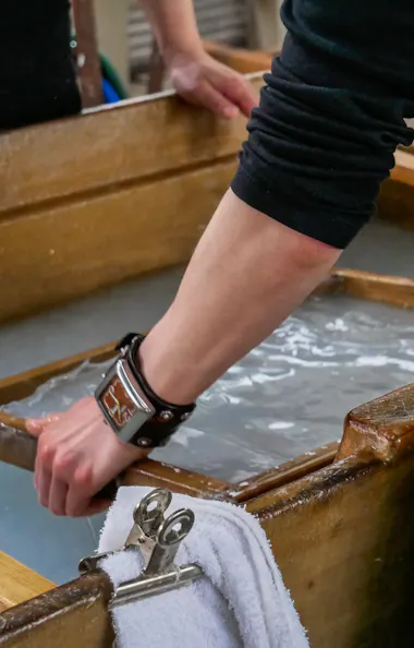 Washi Papermaking A person wearing a leather wristband uses a wooden frame to make handmade paper in a vat of water, with a towel and metal clip clipped to the side of the wooden container.