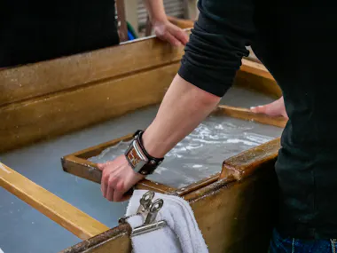 A person wearing a leather wristband uses a wooden frame to make handmade paper in a vat of water, with a towel and metal clip clipped to the side of the wooden container.