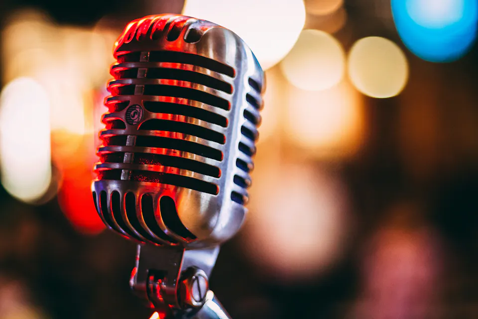 Close-up of a retro-style silver microphone, with colorful bokeh lights in the background. The microphone appears to be set up for a performance or recording, emphasizing its vintage design and the vibrant atmosphere surrounding it.