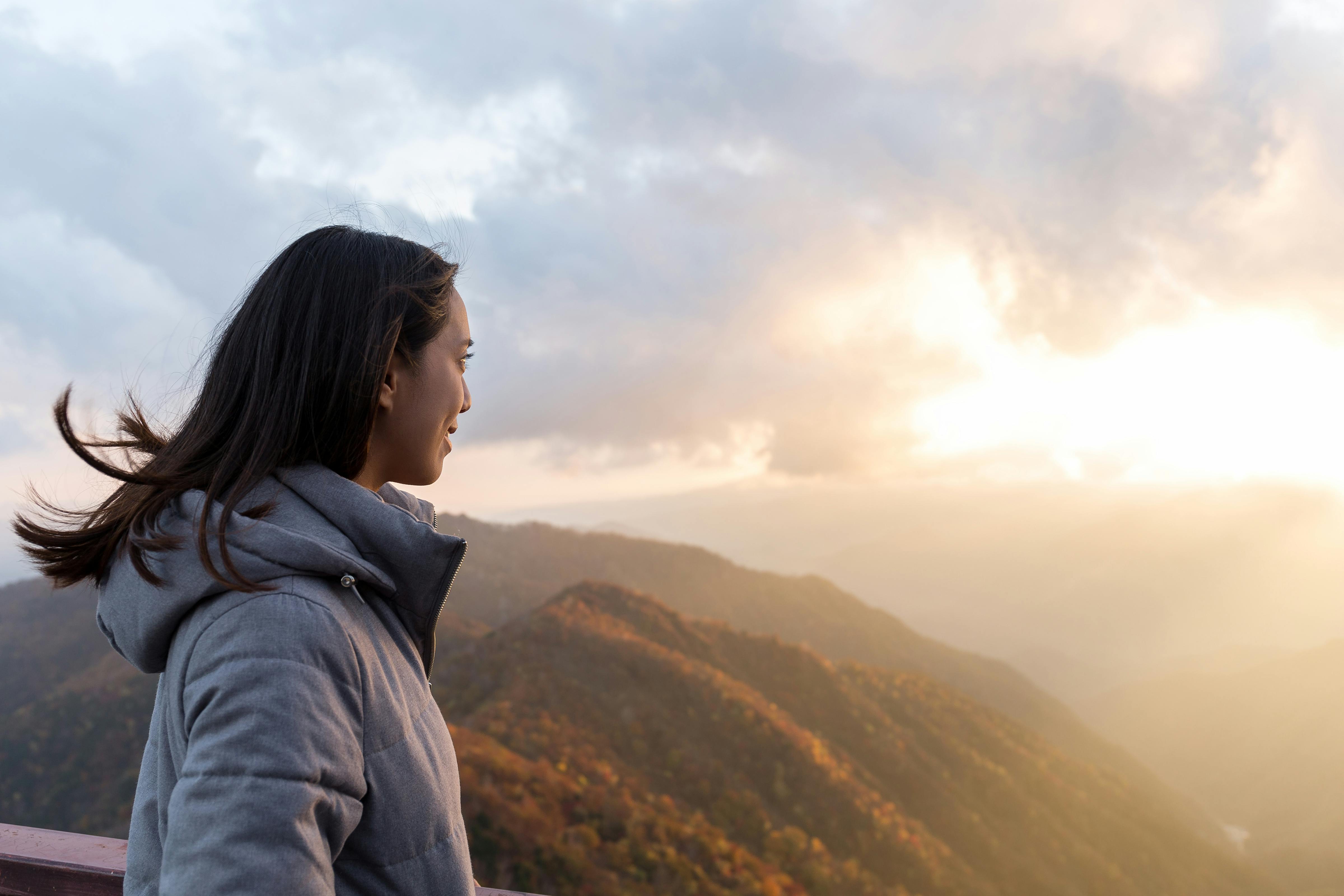 A woman in a gray jacket stands on a balcony, looking out over sunlit mountains and a cloudy sky at sunrise or sunset.
