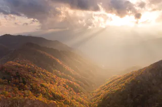 Sunlight streams through clouds, casting warm rays over rolling forested hills covered in autumn foliage, with shades of orange, yellow, and green visible in the landscape below.