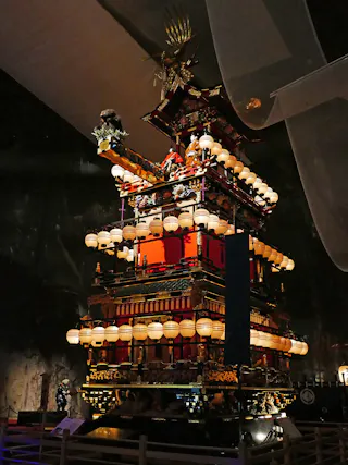 A traditional Japanese festival float, elaborately decorated with red panels, gold accents, and many illuminated lanterns, stands on display in a dimly lit indoor setting.
