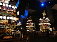Illuminated traditional Japanese festival floats with ornate decorations and lanterns are displayed indoors. A person stands nearby, observing the vibrant structures under dramatic lighting.