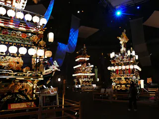 Illuminated traditional Japanese festival floats with ornate decorations and lanterns are displayed indoors. A person stands nearby, observing the vibrant structures under dramatic lighting.