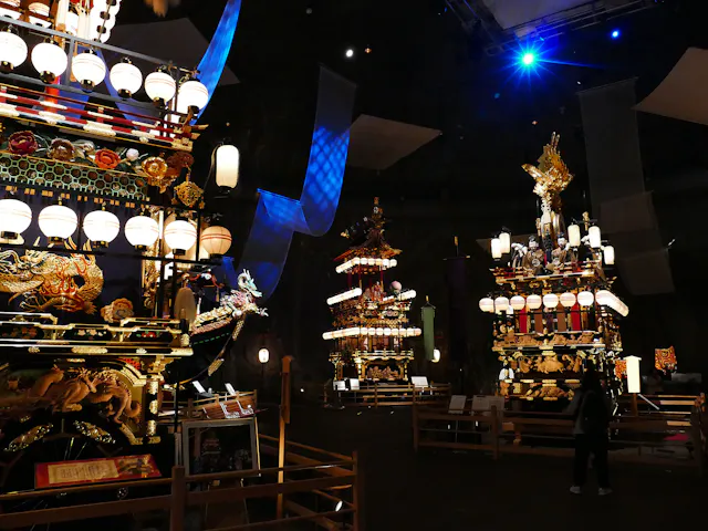 Illuminated traditional Japanese festival floats with ornate decorations and lanterns are displayed indoors. A person stands nearby, observing the vibrant structures under dramatic lighting.